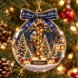 Könnte beinhalten: Ein runder, transparenter Acryl-Weihnachtsanhänger mit einer blauen Schleife oben und der Aufschrift Merry Christmas 2026. Im Inneren klettert ein Arbeiter auf einen Mast inmitten von verschneiten Tannen, Laternen und Geschenken.