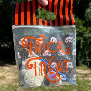 May include: A clear plastic bag with an orange and black striped top containing a white ceramic jar labeled WORM WORT, a blue paintbrush, and small paint pots. The bag features a black background with a skull pattern and orange text reading TRICK or TREAT.
