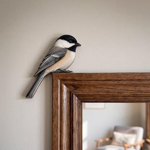 May include: A black-capped chickadee figurine with a black head, white cheeks, and grey wings perched on the top corner of a rectangular wooden mirror frame. The bird has a small black beak and a long tail, resting against a plain wall surface.