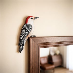 May include: A carved wooden woodpecker figurine featuring a bright red crown, a white face, and black and white barred wings. The bird is perched on the top corner of a dark brown wooden mirror frame, displaying detailed feather textures.
