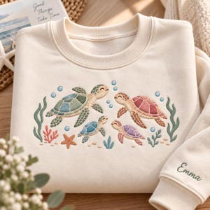May include: A cream-coloured crewneck sweatshirt featuring an embroidered design of four sea turtles in shades of green, red, blue, and purple, surrounded by coral, seaweed, and bubbles. The name Emma is embroidered in green on the sleeve cuff.