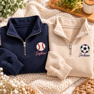 Può includere: Una felpa blu navy con zip a un quarto, un baseball ricamato e il nome Sophia in rosa, accanto a una felpa beige con zip a un quarto, un pallone da calcio ricamato e il nome Jackson in blu navy, entrambe dotate di cerniere color argento.