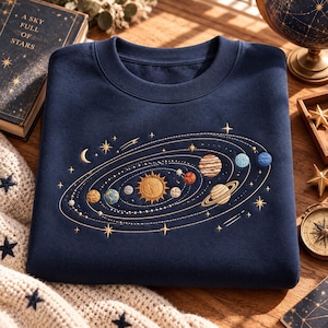Könnte beinhalten: Ein marineblaues Sweatshirt mit Rundhalsausschnitt und einer detaillierten Stickerei des Sonnensystems. Das Design zeigt die Sonne in der Mitte, umgeben von verschiedenen Planeten, Monden und Sternen in Gold-, Blau- und Brauntönen.