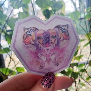 Pode incluir: Um autocolante em forma de coração com uma ilustração surrealista. Dois tigres emolduram uma imagem central de uma mulher com rostos divididos. A obra de arte é predominantemente rosa e roxa, com detalhes intrincados e um tema místico.