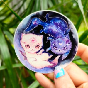 May include: A round sticker featuring three stylized cat illustrations. One cat is white with a sun symbol, another is purple with a crescent moon, and the third is a blue cat-shaped nebula. The background is dark with a starry design.