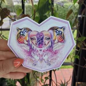 Pode incluir: Um autocolante em forma de coração com uma ilustração detalhada. Dois tigres ladeiam uma imagem central de uma mulher com rostos divididos, um com um design de galáxia. A obra de arte é em tons de rosa, roxo e dourado, com a assinatura do artista na parte inferior.