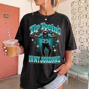 May include: A black short-sleeve t-shirt featuring a graphic of a football player wearing a teal jersey with the number 33. The text "TIP TOEING IN MY JORDANS" is printed in teal above and below the player, surrounded by small star icons.
