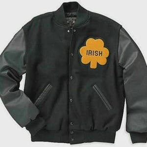 Könnte beinhalten: Eine schwarze Collegejacke mit langen Ärmeln, einer Druckknopfleiste auf der Vorderseite und zwei Seitentaschen. Auf der linken Brust befindet sich ein großer, gelber, kleeblattförmiger Aufnäher mit dem in Schwarz gestickten Wort IRISH.