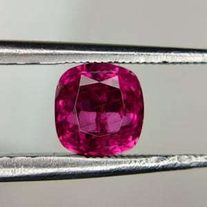 Puede incluir: Una piedra preciosa de corte cojín con un color magenta rosado intenso y vibrante, con una superficie facetada. La piedra está sujeta entre dos puntas metálicas, mostrando su calidad translúcida y su corte geométrico de caras pulidas.