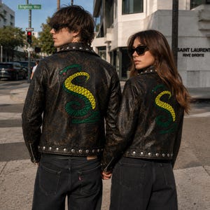 May include: Two black leather motorcycle jackets featuring a large, embroidered snake graphic on the back. The snake is coiled in an S-shape with green and yellow scales and a red tongue. The jackets have silver metal studs along the bottom hem.