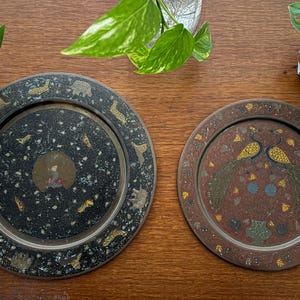 May include: Two circular metal decorative plates. The left plate is black with gold-toned animal silhouettes and a central circular motif. The right plate is brown with a central illustration of two peacocks and floral patterns in gold and blue.