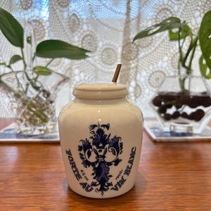 May include: A white ceramic jar with a lid and a small wooden spoon. The jar features a blue floral design and the text "FORTE AU VIN BLANC" with "6 1/2 oz" and "190g" printed on the front. The container has a rounded body and a short neck.