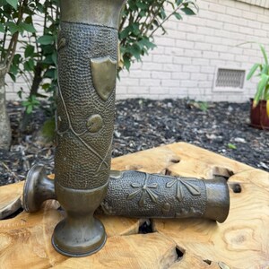 May include: Two cylindrical brass vases with a dark, aged patina. Each vase features a textured, hammered surface with embossed floral motifs and a smooth shield-shaped emblem. The vases have a flared rim and a circular base for stability.