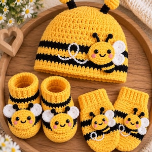 May include: A yellow and black striped crochet hat, a pair of matching booties, and a pair of mittens. Each item features a small, smiling bee appliqué with white wings, black antennae, and pink cheeks. The hat includes two small yellow antennae on top.