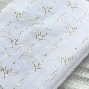 May include: White tissue paper featuring a repeating pattern of the gold-colored logo KM and the text KINDREDMARK STUDIO, interspersed with small gold heart icons. The paper has a subtle, light gray marble-like texture throughout the surface.