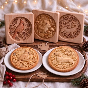 May include: Three square wooden cookie molds featuring carved designs of a cardinal, a hedgehog, and a dachshund, accompanied by two round cookies embossed with the cardinal and dachshund designs, all displayed on a wooden tray.