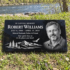 May include: A rectangular black granite memorial plaque featuring an engraved portrait of a man, the text "IN LOVING MEMORY ROBERT WILLIAMS JULY 6, 1960 – APRIL 15, 2025 Beloved Husband, Father & Friend Your spirit lives on in the places you loved.", and a mountain scene with an eagle.