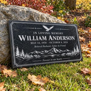 May include: A rectangular black granite memorial plaque featuring an engraved eagle, mountain range, and pine forest. The text reads: IN LOVING MEMORY, WILLIAM ANDERSON, MAY 14, 1958 - OCTOBER 8, 2024, Beloved Husband, Father & Friend, Forever in our hearts.