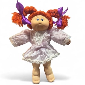 May include: A doll with orange yarn hair styled in two pigtails tied with purple ribbons. The doll has blue eyes and a round face, wearing a light purple dress with white lace trim, a V-neck bodice with three buttons, and a tied waist sash.