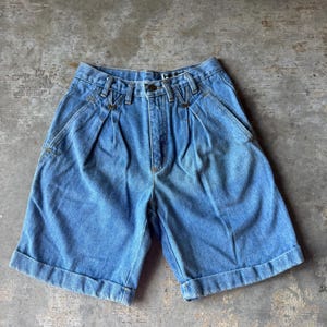 May include: Light blue denim shorts featuring a high-waisted design with double front pleats, a button and zipper fly closure, belt loops, and cuffed hems. The shorts have a classic five-pocket style construction with visible gold-tone stitching.