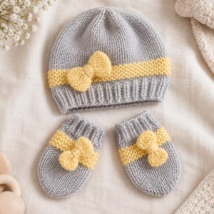 May include: A light gray knitted beanie hat and a matching pair of mittens. Each piece features a horizontal band of yellow garter stitch texture and a small, centered yellow knitted bow detail on the front of the hat and each mitten.
