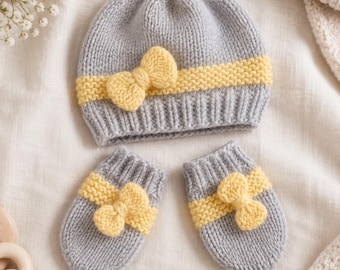 May include: A light grey knitted beanie hat and a matching pair of mittens. Each piece features a horizontal band of yellow garter stitch texture and a small, centred yellow knitted bow detail on the front of the hat and each mitten.