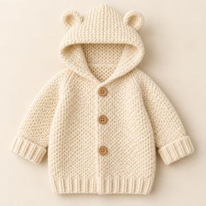 May include: A cream-colored, long-sleeved knit cardigan featuring a hood with two small, rounded bear ears on top. The garment is fastened down the front with three light brown, four-hole wooden buttons and has ribbed cuffs and a ribbed hem.
