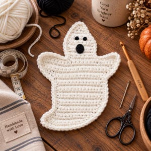 May include: A white crocheted ghost appliqué with two small black circular eyes and one larger black circular mouth. The ghost is positioned among crafting supplies including a wooden 5.0 mm crochet hook, scissors, and a measuring tape.