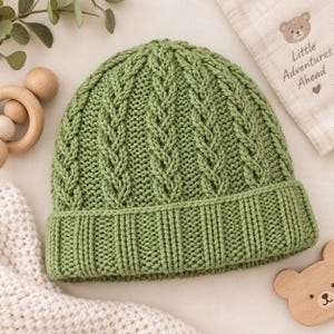 May include: A sage green knitted beanie featuring a vertical cable knit pattern and a wide ribbed cuff. The hat is constructed with a thick yarn texture and displays a consistent stitch throughout the crown and the folded brim section.
