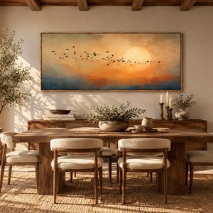 Könnte beinhalten: Ein rechteckiges Landschaftsgemälde, das einen Vogelschwarm zeigt, der durch einen Sonnenuntergangshimmel mit orangefarbenen, gelben und blauen Tönen fliegt. Das Kunstwerk hängt an einer hellen Wand über einem hölzernen Esstisch mit Stühlen.