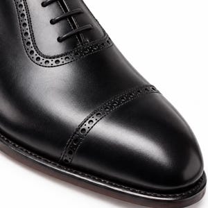 Close-up of black leather cap toe Oxford shoe showing polished finish, decorative brogue perforations and refined formal toe shape.