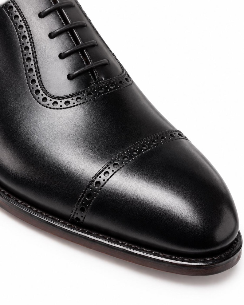 Close-up of black leather cap toe Oxford shoe showing polished finish, decorative brogue perforations and refined formal toe shape.