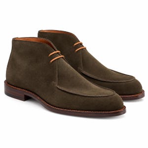 Op de afbeelding: Een paar olijfgroene suède chukka-laarzen met een mocassin-stijl gestikte neus, twee oogjes met dunne lichtbruine veters en een donkerbruine gestapelde leren hak en zool met zichtbare stiksels langs de rand van de schoenzool.