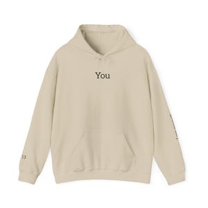 Puede incluir: Una sudadera con capucha de color beige que cuenta con un bolsillo tipo canguro frontal y mangas largas. La palabra "You" está impresa en negro en el centro del pecho. El número "13" aparece en el puño derecho y "Love yourself" en la manga izquierda.