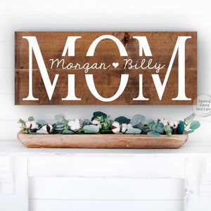Op de afbeelding: Een houten bord met het woord "MOM" in grote witte letters. De namen "Morgan" en "Billy" staan in kleinere witte letters onder het woord "MOM".