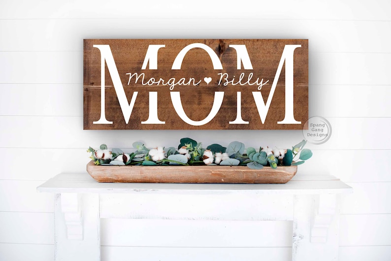 Op de afbeelding: Een houten bord met het woord "MOM" in grote witte letters. De namen "Morgan" en "Billy" staan in kleinere witte letters onder het woord "MOM".