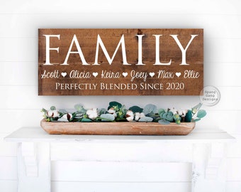 May include: A brown wooden sign with the word "FAMILY" in white lettering. Below the word "FAMILY" are the names "Scott, Alicia, Keira, Joey, Max, Ellie" with a heart symbol between each name. The sign also includes the text "Perfectly Blended Since 2020".