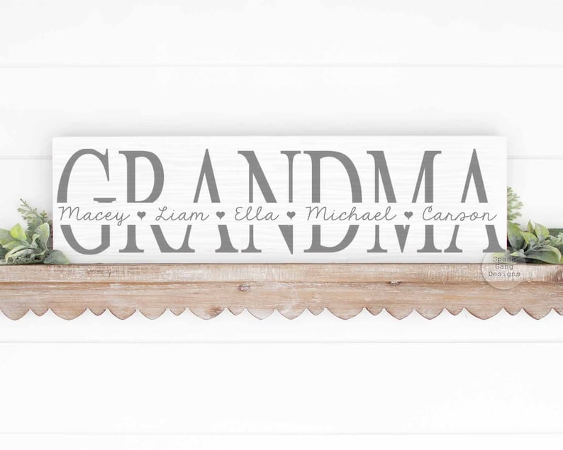 Op de afbeelding: Een wit houten bord met het woord "Grandma" in grijze letters. Onder het woord "Grandma" staan de namen "Macey", "Liam", "Ella", "Michael" en "Carson" met een hartje tussen elke naam.