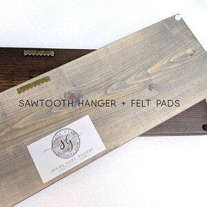 May include: A gray wooden sign with a sawtooth hanger and felt pads on the back. The sign has a white label with the text "Spang Gang Designs" and a logo.