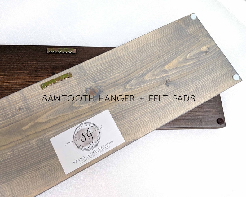 May include: A gray wooden sign with a sawtooth hanger and felt pads on the back. The sign has a white label with the text "Spang Gang Designs" and a logo.