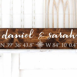 May include: A dark brown wooden sign with white text that reads "daniel & sarah" and coordinates "N 39° 36′ 43.8" W 84° 10' 0.4". A compass rose is centred between the names and coordinates.