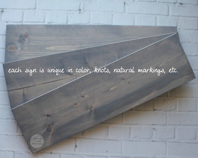 May include: Three pieces of wood, stacked on top of each other, with a gray stain. The wood has a natural grain and knots. The text "each sign is unique in color, knots, natural markings, etc." is written on the wood. The text "Spang Gang Designs" is written on a small circle in the bottom right corner.
