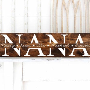 Op de afbeelding: Een bruin houten bord met het woord "NANA" in witte letters. Onder het woord "NANA" staan de namen "Macey", "Liam", "Ella", "Michael" en "Carson" in witte cursieve letters, gescheiden door kleine witte hartjes.