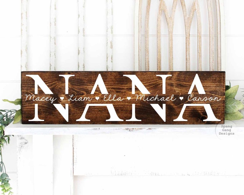 Op de afbeelding: Een bruin houten bord met het woord "NANA" in witte letters. Onder het woord "NANA" staan de namen "Macey", "Liam", "Ella", "Michael" en "Carson" in witte cursieve letters, gescheiden door kleine witte hartjes.