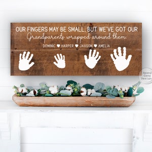 May include: Wooden sign with the text "Our fingers may be small, but we've got our grandparents wrapped around them."  The sign also includes four handprints and the names Dominic, Harper, Jaxson, and Amelia.