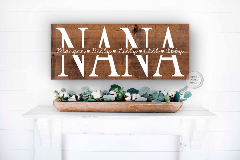 Op de afbeelding: Een houten bord met het woord "NANA" in grote witte letters. Onder het woord "NANA" staat een kleinere regel tekst die luidt "Morgan &hearts; Billy &hearts; Lilly &hearts; Will &hearts; Abby".
