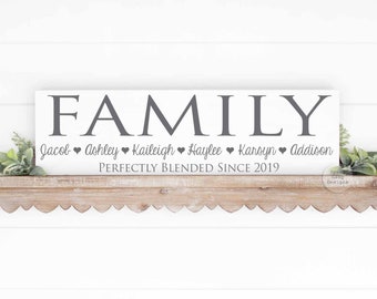 May include: A white wooden sign with the word "FAMILY" in large grey letters. Below the word "FAMILY" are the names Jacob, Ashley, Kaileigh, Haylee, Karsyn, and Addison, each followed by a heart symbol. The text "Perfectly Blended Since 2019" is below the names.