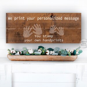 May include: A wooden sign with the text "We print your personalized message. You stamp your own handprints."  The sign has three handprint outlines in white. The sign is hanging on a white wall.