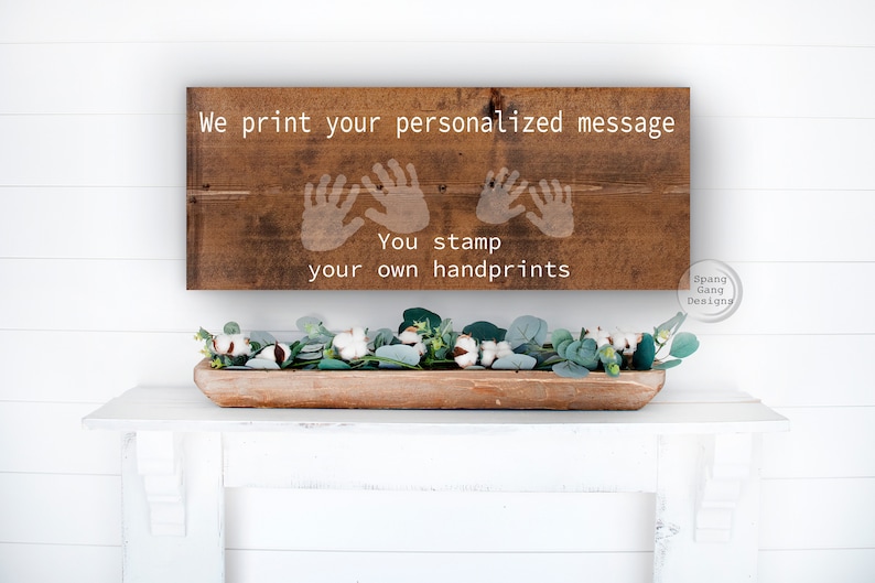 May include: A wooden sign with the text "We print your personalized message. You stamp your own handprints."  The sign has three handprint outlines in white. The sign is hanging on a white wall.