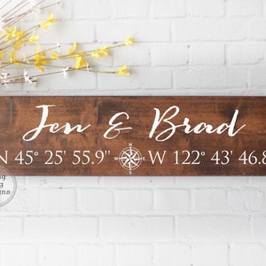 May include: A wooden sign with the names "Jen & Brad" and coordinates "N 45&deg; 25' 55.9" W 122&deg; 43' 46.8" written in white paint. A compass rose is also included on the sign.